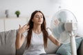 Sad young european woman suffers from unbearably too hot weather, catches cold air from fan in living room