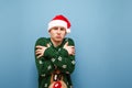 Sad young man in green warm sweater and santa hat warms up against blue background,guy freezes, looks in camera with sad face. Royalty Free Stock Photo
