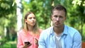 Sad young man looking camera, annoyed jealous girlfriend holding phone, distrust Royalty Free Stock Photo