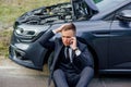 Sad young man use smartphone sitting on road near the broken car opened the hood, calling car assistance services Royalty Free Stock Photo