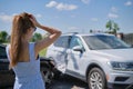 Sad young woman driver standing near her smashed car looking shocked on crashed vehicles in road accident