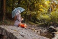 Sad young woman sitting on bringe by pond in autumn park under transparent umbrella during rain. Fall season walk Royalty Free Stock Photo