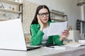 Sad young woman working from home with laptop and documents. Holds in hands and anxiously looks at bills, letter