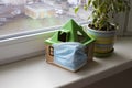 Safe home concept, quarantine coronavirus. a toy house in a medical mask stands on a windowsill next to a flower in a Royalty Free Stock Photo
