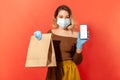 Safe online shopping on quarantine. Woman in protective mask and gloves holding package with food and mobile phone Royalty Free Stock Photo