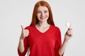 Safe pleaure and protection concept. Glad redhaired female with freckled skin holds condom, shows ok sign, dressed in red clothes, Royalty Free Stock Photo