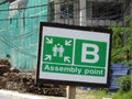 Safety signage `Emergency Assembly Point` with direction arrow at the construction site.