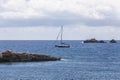 Sailing boat without sails maneuvers between rocks protruding above shallow water under heavy low white clouds. Ibiza Royalty Free Stock Photo