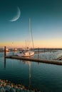 a sailing harbor there is a white sailboat in the sunset and in the sky there is the big half moon Royalty Free Stock Photo