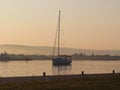 The sailing yacht enters under the motor in the marina in the rays of the morning sun. Dawn in the port. Empty pier with electrica