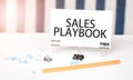 SALES PLAYBOOK sign on paper on white desk with office tools. Blue and white background