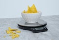 Salted tortilla nacho chips in a white bowl on a kitchen scale, grey marble table