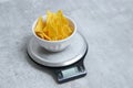 Salted tortilla nacho chips in a white bowl on a kitchen scale, grey marble table