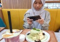 Samarinda, Indonesia. January 23th 2023. Happy Asian muslim teenage girl sitting in cafe and taking picture of her food with smart