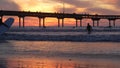 People surfing, pier on piles at sunset. Surfer in ocean water waves, California