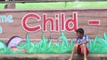 Boy sitting on sidewalk pavement of Mural painted concrete school wall waiting for his playmates.