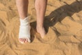 Male right leg in white cast and bandages closeup. Left foot immersed in sand