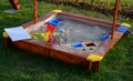 Children`s small wooden sandpit with plastic toys around the lawn brown painted yellow corners green red blue Royalty Free Stock Photo