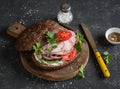 Sandwich with roasted pork, soft cheese, tomatoes and arugula on the wooden cutting board on dark background. Royalty Free Stock Photo