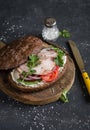 Sandwich with roasted pork, soft cheese, tomatoes and arugula on the wooden cutting board on dark background. Royalty Free Stock Photo