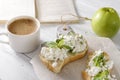 Sandwiches with soft cheese and Basil on a plate, a Cup of hot black coffee, a green Apple, a book on a white table, a delicious