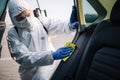 Sanitizing service worker cleans the car`s interior with a yellow rug. A man in a protective suit, mask and gloves disinfects the