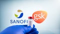 Sanofi and gsk logo and covid-19 coronavaccine injection vial held with blue medical gloves.