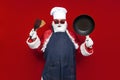 santa claus in chef\'s hat and apron holds frying pan and utensils on red background, male cook in santa costume Royalty Free Stock Photo