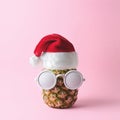 Santa Claus face made from pineapple and white painted sunglasses on pink background.