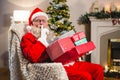 Santa claus with gift boxs sitting on chair with finger on lips in living room