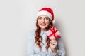 Santa Claus girl with curly hair and a red hat with a bumbon holds a gift box with a red satin ribbon bow and smiles on a