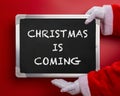 Santa Claus holding a black chalk board written with CHRISTMAS IS COMING on red Royalty Free Stock Photo