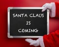 Santa Claus holding a black chalk board written with SANTA CLAUS IS COMING on red Royalty Free Stock Photo