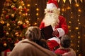 Santa Claus reading book for family. Mother and children sitting indoor near decorated xmas tree with lights - Merry Christmas and Royalty Free Stock Photo