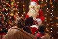Santa Claus reading book for family. Mother and children sitting indoor near decorated xmas tree with lights - Merry Christmas and Royalty Free Stock Photo