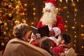 Santa Claus reading book for family. Mother and children sitting indoor near decorated xmas tree with lights - Merry Christmas and Royalty Free Stock Photo