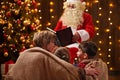 Santa Claus reading book for family. Mother and children sitting indoor near decorated xmas tree with lights - Merry Christmas and Royalty Free Stock Photo