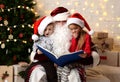 Santa Claus reading magic book to happy little cute children boy and girl kids near Christmas tree Royalty Free Stock Photo