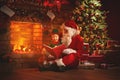 Santa claus reads a book to a little elf by Christmas tree