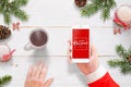 Santa Claus send Merry Christmas message with mobile phone. Hot tea, and Christmas decoration beside