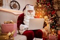 Santa Claus sitting at his home and reading email on laptop with ÃÂhristmas requesting or wish list near the fireplace and tree Royalty Free Stock Photo