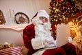 Santa Claus sitting at his home and reading email on laptop with ÃÂhristmas requesting or wish list near the fireplace and tree Royalty Free Stock Photo