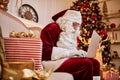 Santa Claus sitting at his home and reading email on laptop with ÃÂhristmas requesting or wish list near the fireplace and tree Royalty Free Stock Photo