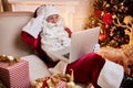 Santa Claus sitting at his home and reading email on laptop with ÃÂhristmas requesting or wish list near the fireplace and tree Royalty Free Stock Photo