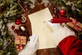 Santa Claus writing with a vintage quill pen on a sheet of blank paper surrounded by pine foliage and Christmas decorations viewed