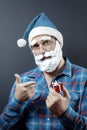 Santa with a foam beard holds a tiny red gift box and points his index finger at it. Hand with a gift is defocused