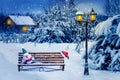 Santa hat and New Year`s soft toy on a bench in the winter forest against the background of a village house and lantern. Christma Royalty Free Stock Photo
