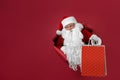 Santa man holds shops packet in hand through a paper hole. Bearded man in santa hat looking through hole on red paper Royalty Free Stock Photo