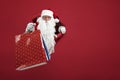 Santa man holds shops packet in hand through a paper hole. Bearded man in santa hat looking through hole on red paper Royalty Free Stock Photo