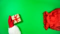 Santa`s hand in a red mitten puts gift boxes wrapped in craft paper and tied with red ribbon into a red bag. Green background Royalty Free Stock Photo